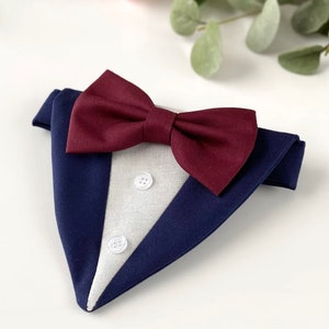 Puede incluir: Un pa&ntilde;uelo triangular para mascotas con dise&ntilde;o de esmoquin, que presenta una base de tela azul marino oscuro, un panel central gris claro, dos peque&ntilde;os botones blancos y una llamativa pajarita color burdeos oscuro de tela texturizada.