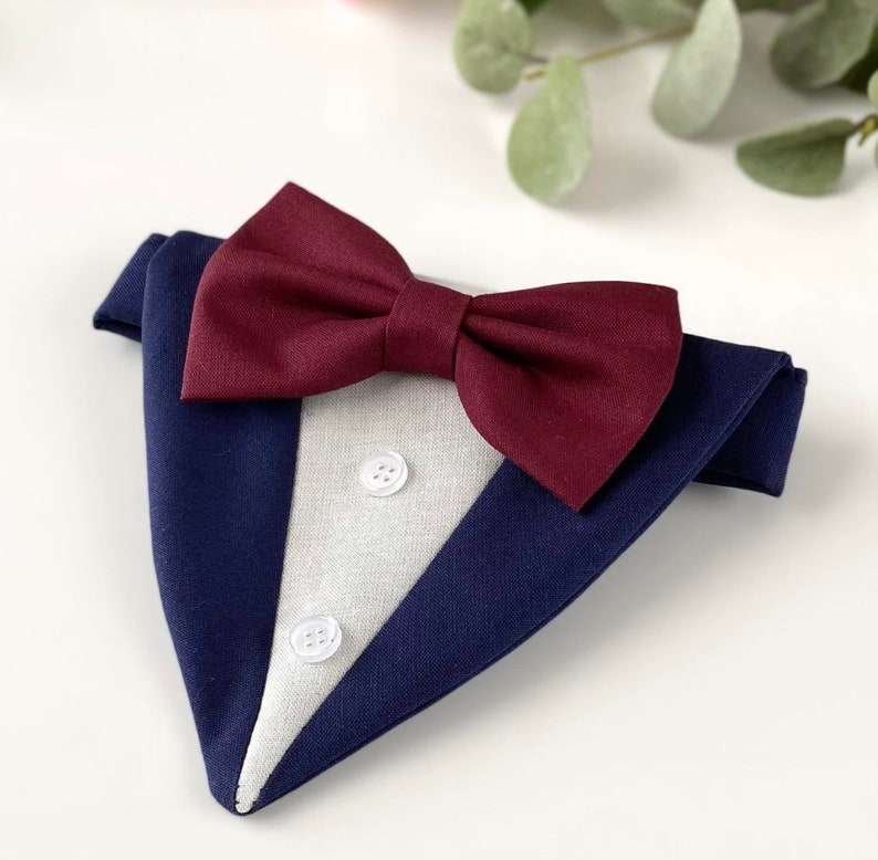 Puede incluir: Un pa&ntilde;uelo triangular para mascotas con dise&ntilde;o de esmoquin, que presenta una base de tela azul marino oscuro, un panel central gris claro, dos peque&ntilde;os botones blancos y una llamativa pajarita color burdeos oscuro de tela texturizada.
