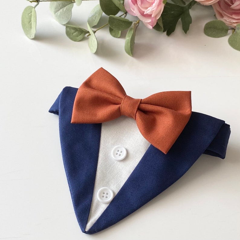 May include: A navy blue fabric pet bandana designed to resemble a tuxedo jacket with a white triangular insert featuring two small white buttons and a prominent burnt orange bow tie attached at the top center of the garment.