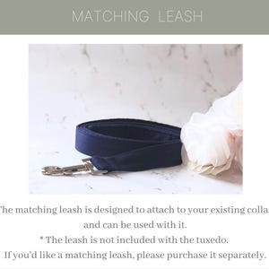 May include: A navy blue fabric dog leash featuring a silver-toned metal swivel snap hook. The leash is coiled and displayed alongside a white floral accent. The text MATCHING LEASH is visible at the top, with additional text below the leash.