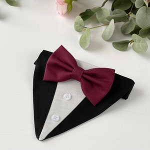 Puede incluir: Un pa&ntilde;uelo triangular para mascotas dise&ntilde;ado como un esmoquin, con tela exterior negra, un panel central gris claro con dos botones blancos y una pajarita de tela color burdeos oscuro sujeta en la parte superior de la prenda.