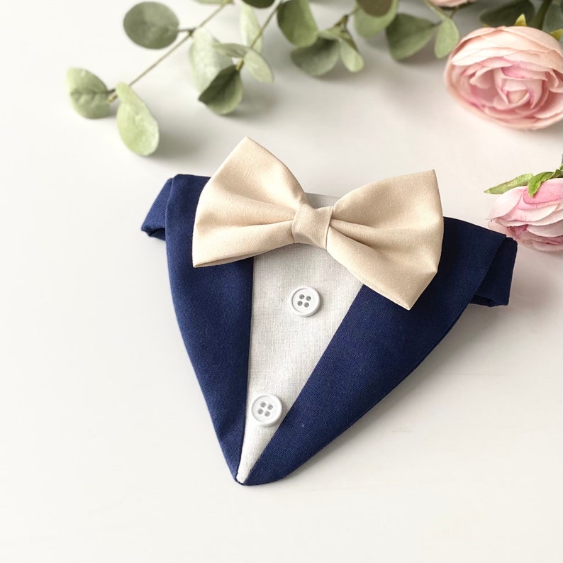 May include: A navy blue tuxedo-style pet bandana featuring a white triangular insert with two small white buttons and a cream-colored bow tie attached at the top. The accessory is designed to mimic a formal suit jacket with a pointed hem.