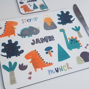 May include: White placemat and coaster set with colorful dinosaur illustrations. The placemat features orange, blue, and green dinosaurs, a volcano, and the name "JAMIE". The coaster has similar designs. Text includes "STOMP" and "MUNCH".