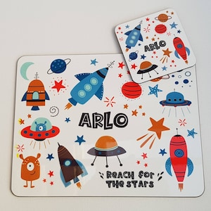 May include: A rectangular placemat and a square coaster featuring a space theme with rockets, planets, stars, and a one-eyed alien. Both items are printed with the name ARLO and the text REACH FOR THE STARS in a bold, dark, sans-serif font.