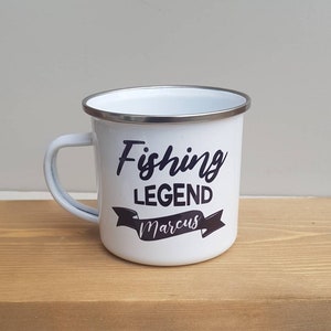 May include: A white enamel mug with a silver-toned rim and a handle on the left side. The front of the mug features black text that reads Fishing LEGEND with the name Marcus printed in a script font inside a black banner graphic below the text.