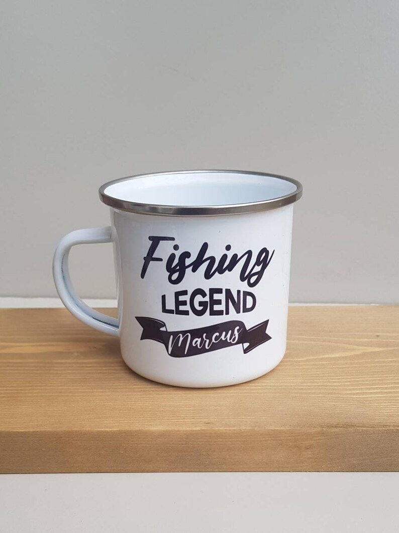 May include: A white enamel mug with a silver-toned rim and a handle on the left side. The front of the mug features black text that reads Fishing LEGEND with the name Marcus printed in a script font inside a black banner graphic below the text.