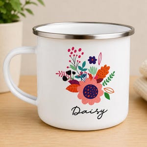 May include: A white enamel mug with a silver rim and a handle on the left side. The front features a colorful floral illustration with pink, orange, green, and dark blue elements. Below the flowers, the name Daisy is printed in black script.