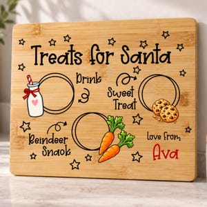 May include: A rectangular bamboo cutting board featuring the text "Treats for Santa" and "love from Ava" in black and red. It includes illustrations of a milk bottle, two chocolate chip cookies, and two carrots, with designated circles for placement.