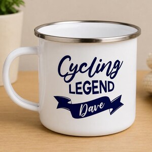 May include: A white enamel mug with a silver-toned rim and a handle on the left side. The front of the mug features dark blue text that reads Cycling LEGEND with the name Dave written in a script font inside a dark blue banner graphic.
