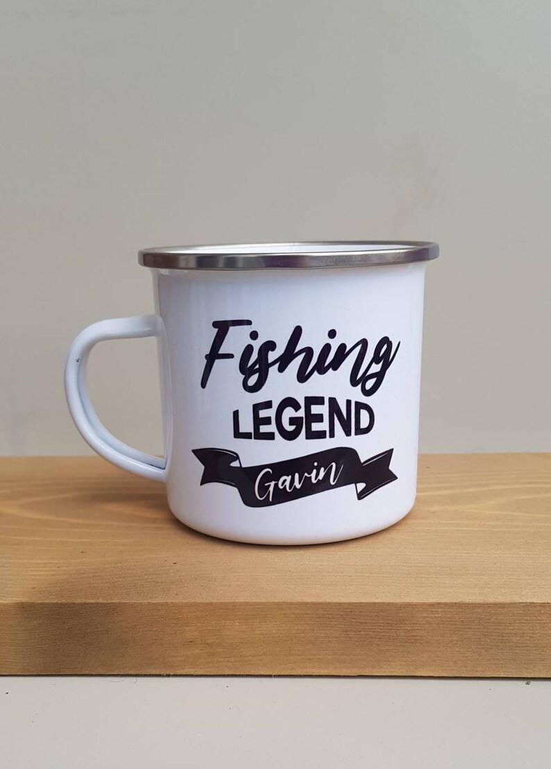 May include: A white enamel mug with a silver rim and a handle. The mug features black text that reads Fishing LEGEND with the name Gavin printed in white script inside a black banner graphic centered on the front of the mug.