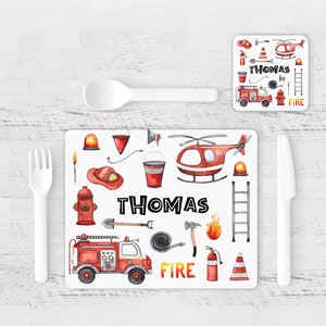 May include: A white placemat and coaster set featuring a firefighter theme. The placemat has a red fire truck, helicopter, and other fire-related illustrations. The name "THOMAS" is printed in black. A white spoon, fork, and knife are also present.