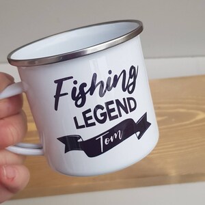 May include: A white enamel mug with a silver-colored rim and a handle. The mug features dark purple text that reads Fishing LEGEND with the name Tom printed inside a dark purple banner graphic below the text.