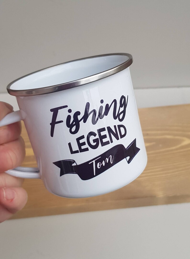 May include: A white enamel mug with a silver-colored rim and a handle. The mug features dark purple text that reads Fishing LEGEND with the name Tom printed inside a dark purple banner graphic below the text.