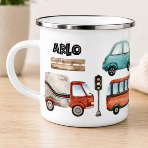 May include: A white enamel mug with a silver rim featuring a watercolor illustration of a teal car, a red truck, a red bus, a traffic light, and a brown road. The name ARLO is printed in black capital letters above the road graphic.