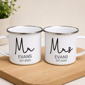 May include: Two white enamel mugs with silver rims. The left mug features black text reading Mr, EVANS, and EST 2020. The right mug features black text reading Mrs, EVANS, and EST 2020. Both mugs have a handle on the side and a smooth finish.