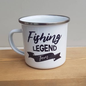 May include: A white enamel mug with a silver-toned rim and a handle on the left side. The front features black text that reads Fishing LEGEND with the name Tom printed in a white script font inside a black banner graphic below the text.
