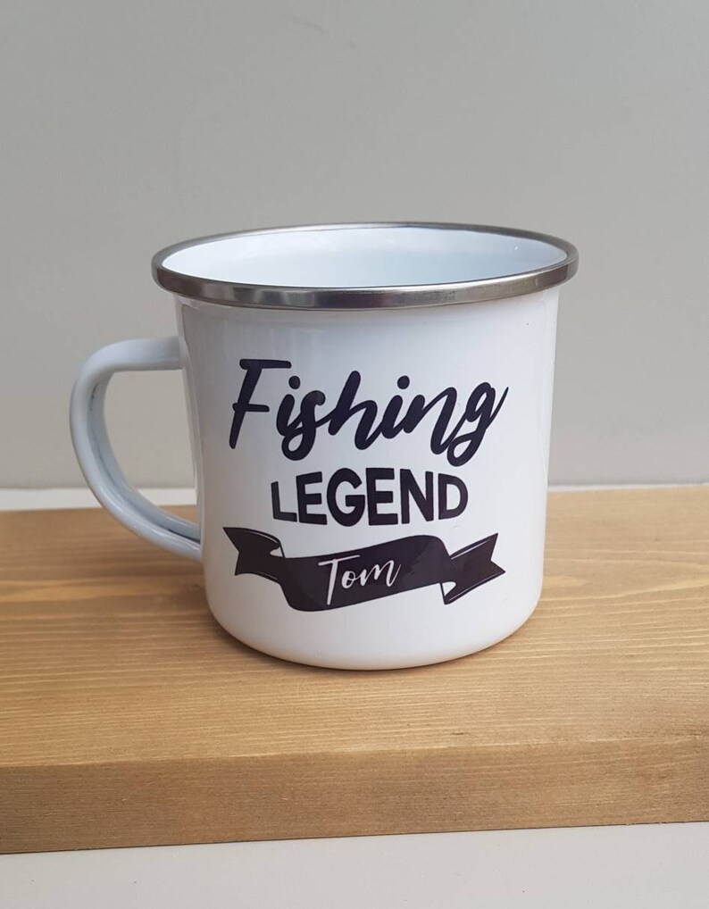 May include: A white enamel mug with a silver-toned rim and a handle on the left side. The front features black text that reads Fishing LEGEND with the name Tom printed in a white script font inside a black banner graphic below the text.