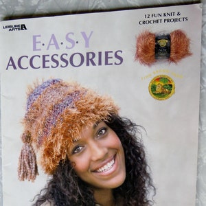 May include: A book cover featuring a woman wearing a brown and purple fuzzy hat with a brown tassel. The book title is "EASY ACCESSORIES" and the cover text says "12 FUN KNIT & CROCHET PROJECTS".