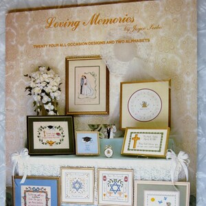 May include: A book cover with the title "Loving Memories" by Joyce Seebo. The cover features a variety of cross-stitch designs in frames, including a wedding scene, a graduation cap, a Star of David, and a bird with a heart.