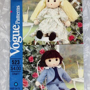 May include: A Vogue Patterns 523 craft envelope featuring two fabric dolls. The top doll has blonde yarn hair, a floral dress with a light green ribbon, and black shoes. The bottom doll has brown yarn hair, a blue suit, and black shoes.