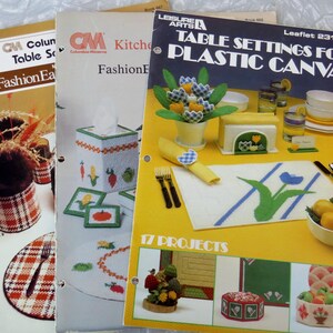 May include: A stack of vintage craft pattern booklets. The covers feature images of table settings, tissue box covers, and other decorative items. The booklet on the right has the title "TABLE SETTINGS FOR PLASTIC CANVAS".