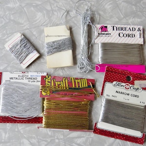 May include: Assortment of metallic threads and cords in silver and gold. Includes spools of elastic metallic thread, narrow cord, and craft trim. Packaging features brand names and product information.