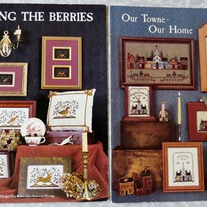 May include: Two craft pattern booklets titled Among the Berries and Our Towne - Our Home. The covers feature photographs of framed cross-stitch designs, including birds, floral motifs, and small town scenes, displayed with decorative home accessories.