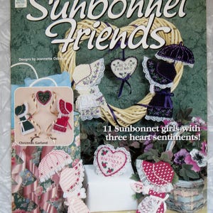 May include: A craft pattern book titled Plastic Canvas Sunbonnet Friends featuring 11 Sunbonnet girls with three heart sentiments. The cover displays various plastic canvas projects including figures in bonnets, heart-shaped ornaments, and a Christmas garland.