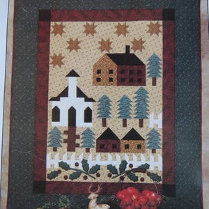 May include: A quilted wall hanging featuring a winter village scene. The design includes a church, houses, trees, and a white picket fence. The background is tan with brown star accents, framed by a dark green border.