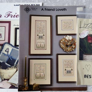 May include: Several cross-stitch patterns and designs are displayed, including framed pieces with text and images. The patterns feature phrases like "Dear Friend" and "Best Friends," along with floral and heart motifs. A gray sweater with a cross-stitch design is also visible.