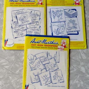 May include: Three yellow paper envelopes containing Aunt Martha's hot iron transfers for embroidery. Each envelope features blue line art illustrations of kitchen-themed designs, including days of the week, dishware, and animated kitchen items.