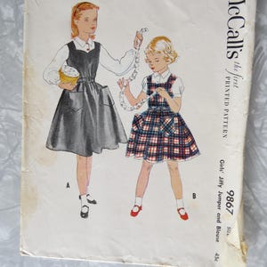 May include: A vintage McCall's printed pattern envelope for a girls' jiffy jumper and blouse, number 9867. The illustration shows two figures wearing jumper dresses with pockets over white collared blouses, one in solid dark gray and one in plaid.