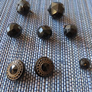 May include: A collection of eight black glass buttons, some with a faceted design and others with a circular design. The buttons are arranged on a blue and white striped surface.