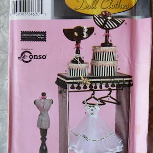 May include: A pink Simplicity Couturier Doll Clothes pattern. The pattern features a doll dress, a display stand, and accessories. The stand has a black and white striped design, with a small dress form and a doll dress on a hanger.