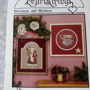 May include: A framed cross-stitch pattern titled "HeartStrings Merriment and Mistletoe." The image features framed cross-stitch art of Santa Claus and an angel, along with decorative elements and text. The price tag reads $0.75.
