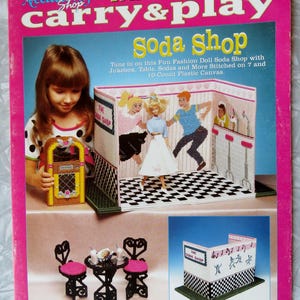 May include: A pink plastic canvas kit titled "Fashion Doll Carry & Play Soda Shop." The cover features a girl with a toy jukebox, and a stitched soda shop scene with a doll, table, and figures. Includes the text "Tune in on this Fun Fashion Doll Soda Shop with Jukebox, Table, Sodas and More Stitched on 7 and 10-Count Plastic Canvas."