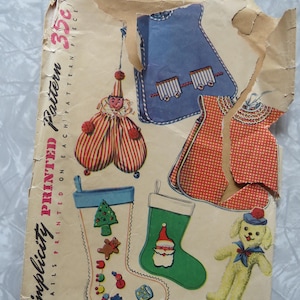 May include: A vintage Simplicity printed pattern envelope featuring illustrations of a clown, a blue bib, a red checkered bib, two Christmas stockings with holiday motifs, and a yellow dog toy. The paper envelope is torn and shows the text 35c.