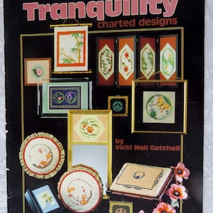 May include: A black cover for a needlework pattern book titled "Tranquility charted designs." The cover features various framed needlework designs with floral and geometric patterns, in shades of green, orange, and yellow. The author's name is "Vicki Neil Getchell."