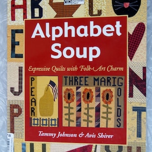 May include: A book cover titled "Alphabet Soup" with the subtitle "Expressive Quilts with Folk-Art Charm." The cover features a patchwork design with large letters, a black cat, and illustrations of a pear and marigolds. The authors are Tammy Johnson & Avis Shirer.