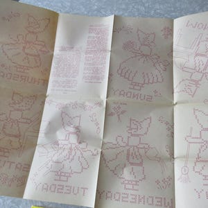 May include: Vintage cross-stitch pattern on aged paper. The pattern features a cat-like figure in various poses, each labelled with a day of the week. The design is in red, with decorative floral elements. The paper is folded.