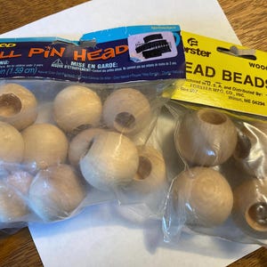 May include: Two clear plastic bags containing light-colored wooden doll pin heads and round beads. The left package is labeled Wood Doll Pin Head 0.625 in (1.59 cm) and contains 10 pieces. The right package is labeled Forster Head Beads.