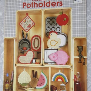 May include: A vintage leaflet titled "Crocheted Potholders" featuring various crochet potholder designs. The designs include a heart, a pig, a watermelon slice, and a rainbow. The leaflet is displayed in a wooden box with a grid background.