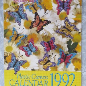May include: A 1992 plastic canvas calendar featuring colorful butterfly designs. The butterflies are in various colors, including blue, pink, yellow, and orange, set against a backdrop of white and yellow flowers. The calendar has the text "Needlecraft Shop".
