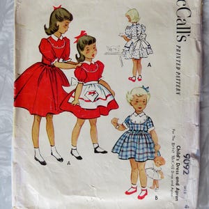 May include: Vintage McCall's sewing pattern for a child's dress and apron. The pattern is for sizes 4 and includes instructions for making a red dress with a white apron and a plaid dress. The pattern is printed on a white background with black text and illustrations.