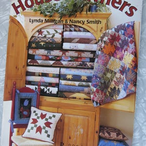 May include: A book titled "House Warmers" by Lynda Milligan & Nancy Smith. The cover features a wooden cabinet filled with colorful quilts and pillows. The book is published by "Possibilities" and features quilt designs, patterns, and fabric suggestions.