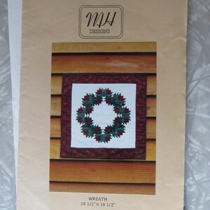 May include: A paper foundation pattern by MH Designs titled Wreath, measuring 18 1/2 inches by 18 1/2 inches. The cover features a square quilt block with a dark red and green wreath design on a white background, set against a wood-paneled wall.