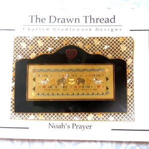 May include: A framed needlework design titled "Noah's Prayer" by The Drawn Thread. The design features zebras, elephants, and other animals. The frame is black with a heart-shaped accent at the top. The background is a patterned fabric.