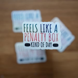 May include: White sticker with the text "Feels like a penalty box kind of day" in black and red lettering. The sticker is on a wooden surface.