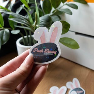 May include: A sticker featuring a bunny head with pink inner ears and a hockey puck design. The text "Puck Bunny BOOKCLUB" is visible on the puck. The sticker is held in a hand, with a plant and white boxes in the background.