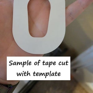 Könnte beinhalten: Ein weißes, ovalförmiges Stück Klebeband mit einem kleineren, ovalförmigen Loch, das aus der Mitte herausgeschnitten ist. Das Klebeband wird in einer Hand gehalten. Der Text "Sample of tape cut with template" ist unterhalb des Klebebands sichtbar.